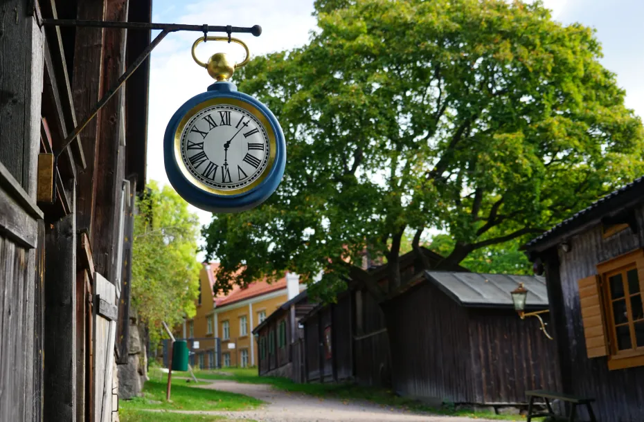 An analog clock hanging from a wooden house in Turku. There are more wooden houses in the background, in a green landscape